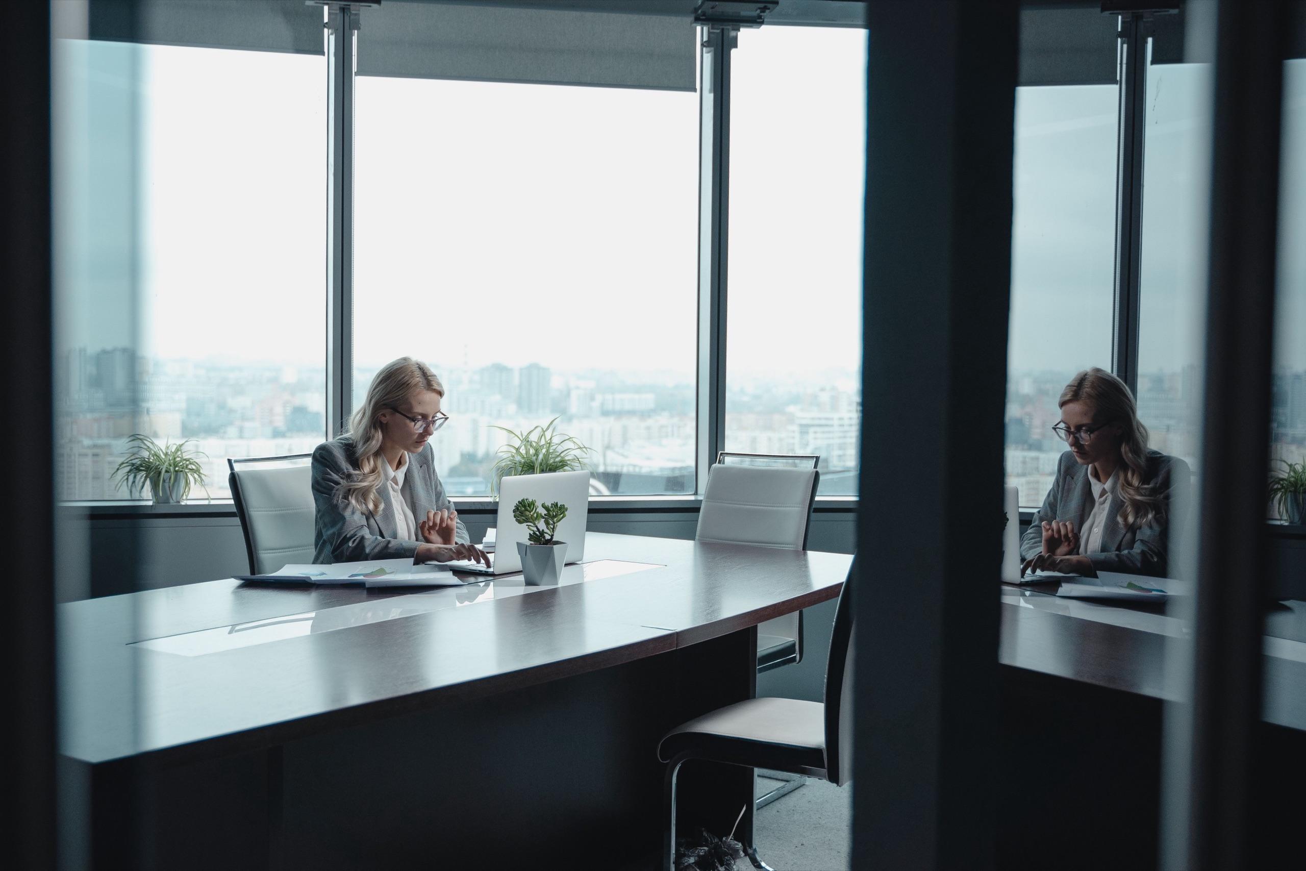 A quiet glass-walled meeting room high above a city, with one person working at a long table beside full-height windows.