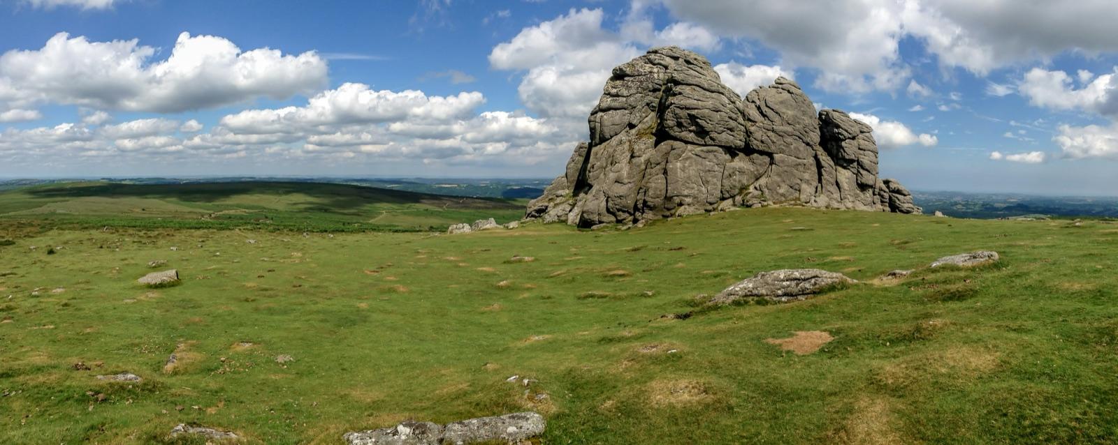 A weathered granite tor standing above open moorland grass, with cumulus cloud building over the hills behind.