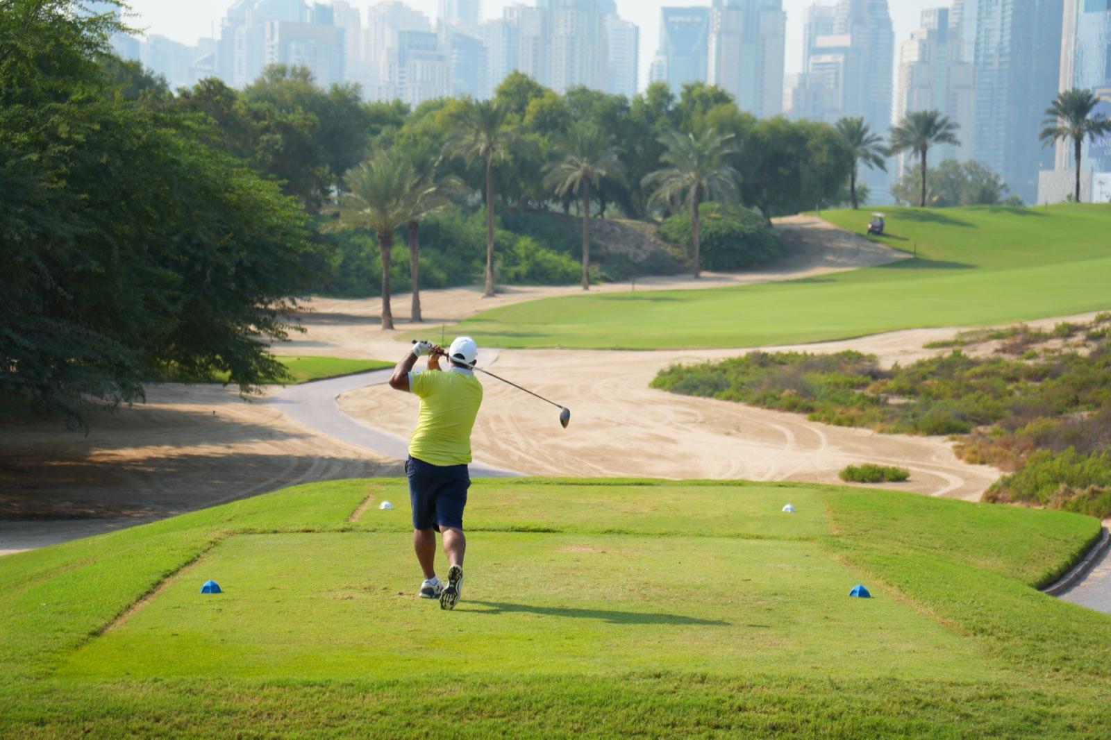 A golfer following through on a tee shot, with palms along the fairway and a high-rise skyline in the haze beyond.