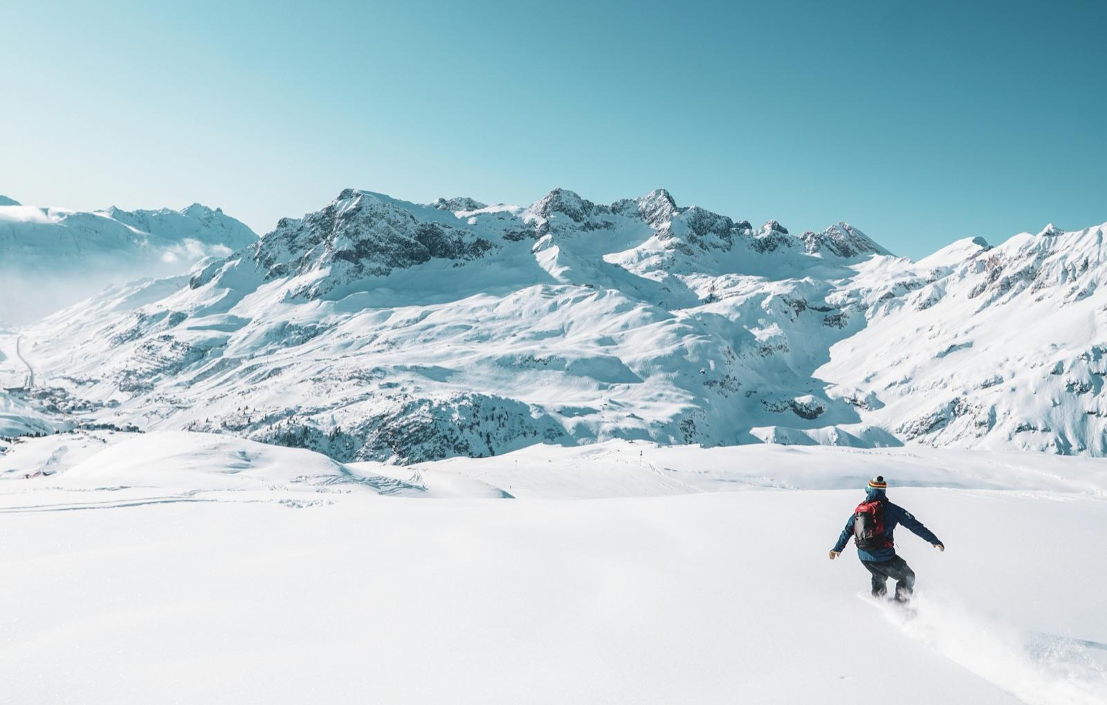 A lone skier crossing an untracked snowfield below a wide range of snow-covered peaks under a clear winter sky.