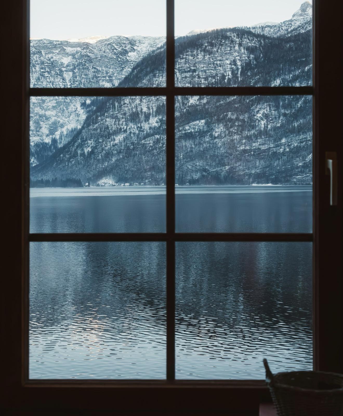 A window with fine glazing bars looking out across a still mountain lake to a snow-covered wooded slope beyond.